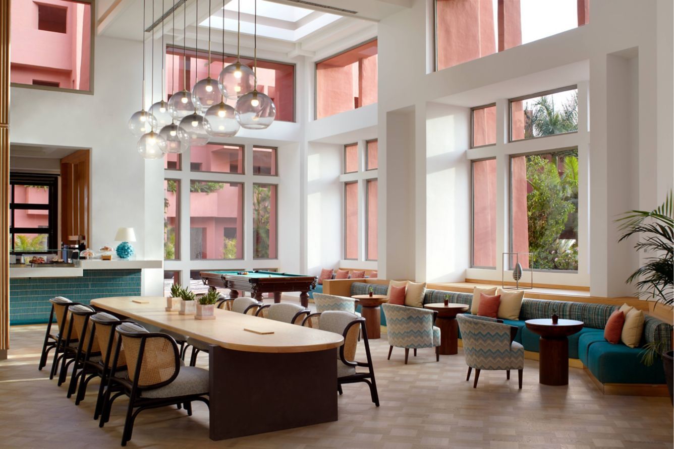 A modern, airy lounge with a long table for dining, soft seating, and large windows in a building with terracotta-colored accents.