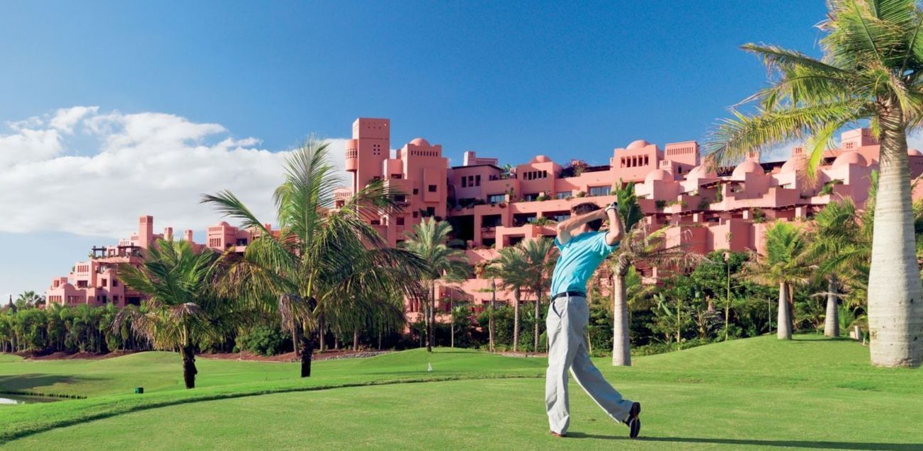 Man in golf attire swings a club on a green course, with a pink resort and palm trees under a blue sky.