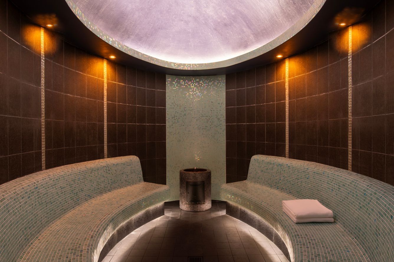 A steam room with curved mosaic tile benches, dark tile walls, and soft lighting. A towel rests on one bench. Spa and Wellness