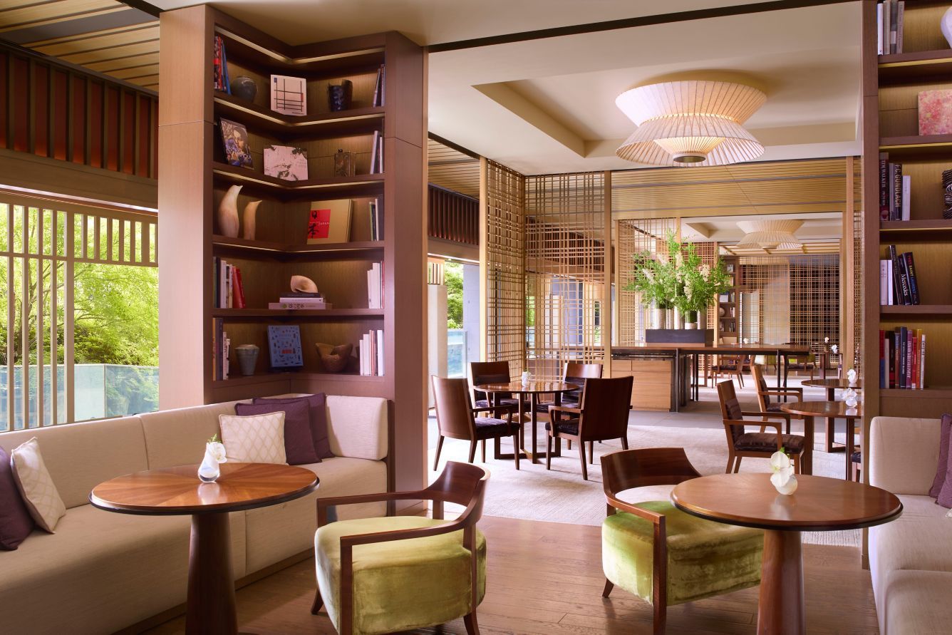 Lounge area with bookshelves, seating, and tables. Beige and brown tones, natural light.