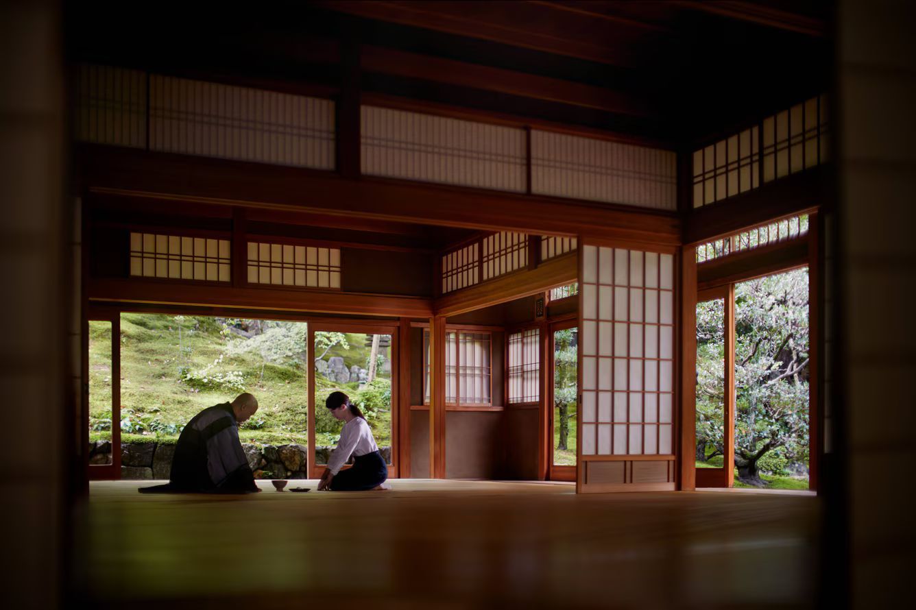 Two people in traditional Japanese room; one bows, the other prepares tea. Wooden structure, garden visible.
