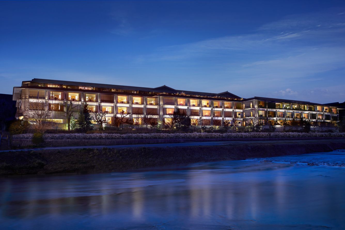 Hotel lit up at night, riverside, with water reflecting the lights. Ritz-Carlton Kyoto