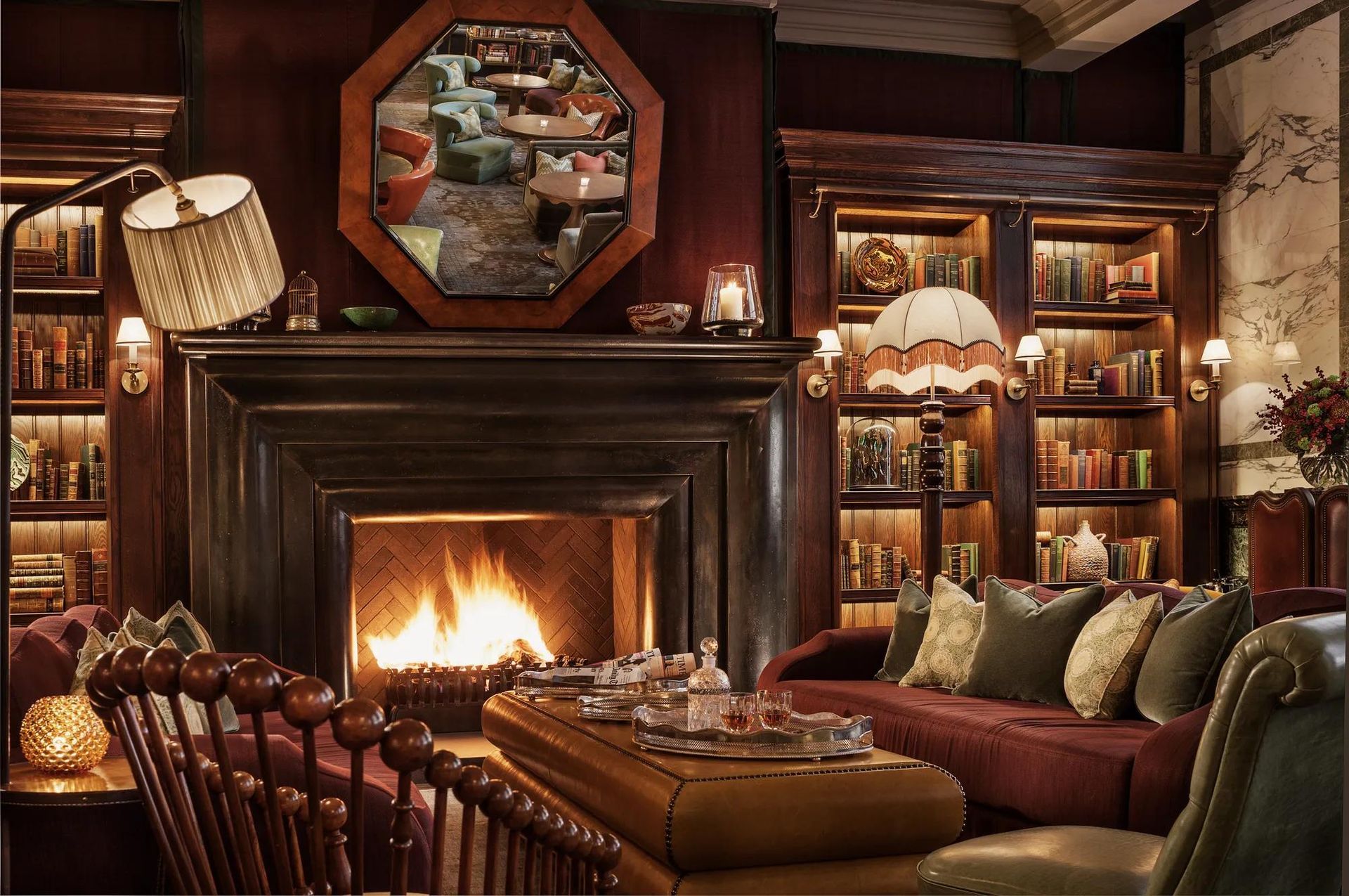 Cozy library lounge with fireplace, bookshelves, and couches in warm red and brown tones. Rosewood London luxury hotel