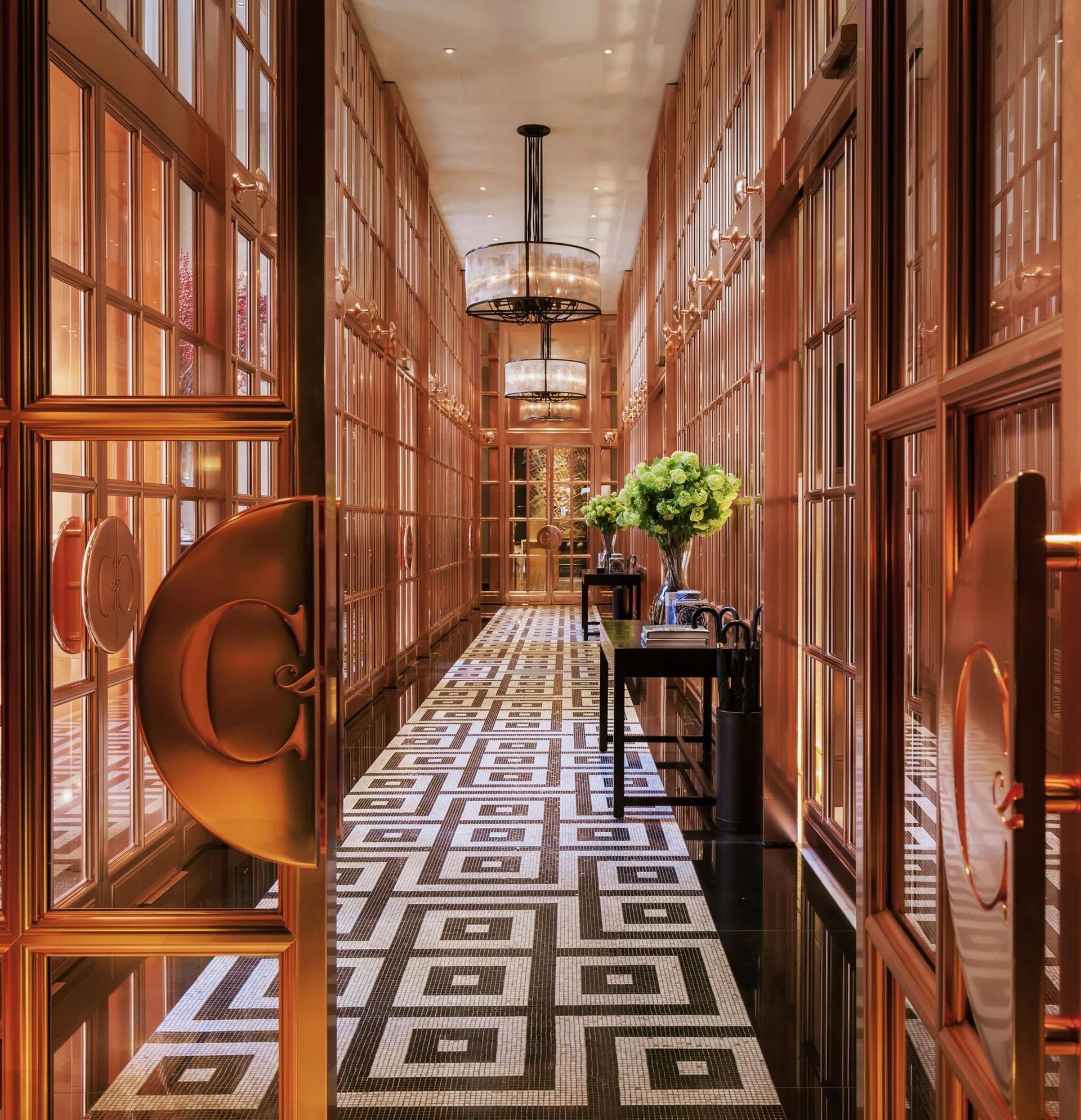 Long hallway with patterned tile floor, glass-paned walls, gold door handles, and chandeliers. Rosewood London luxury hotel