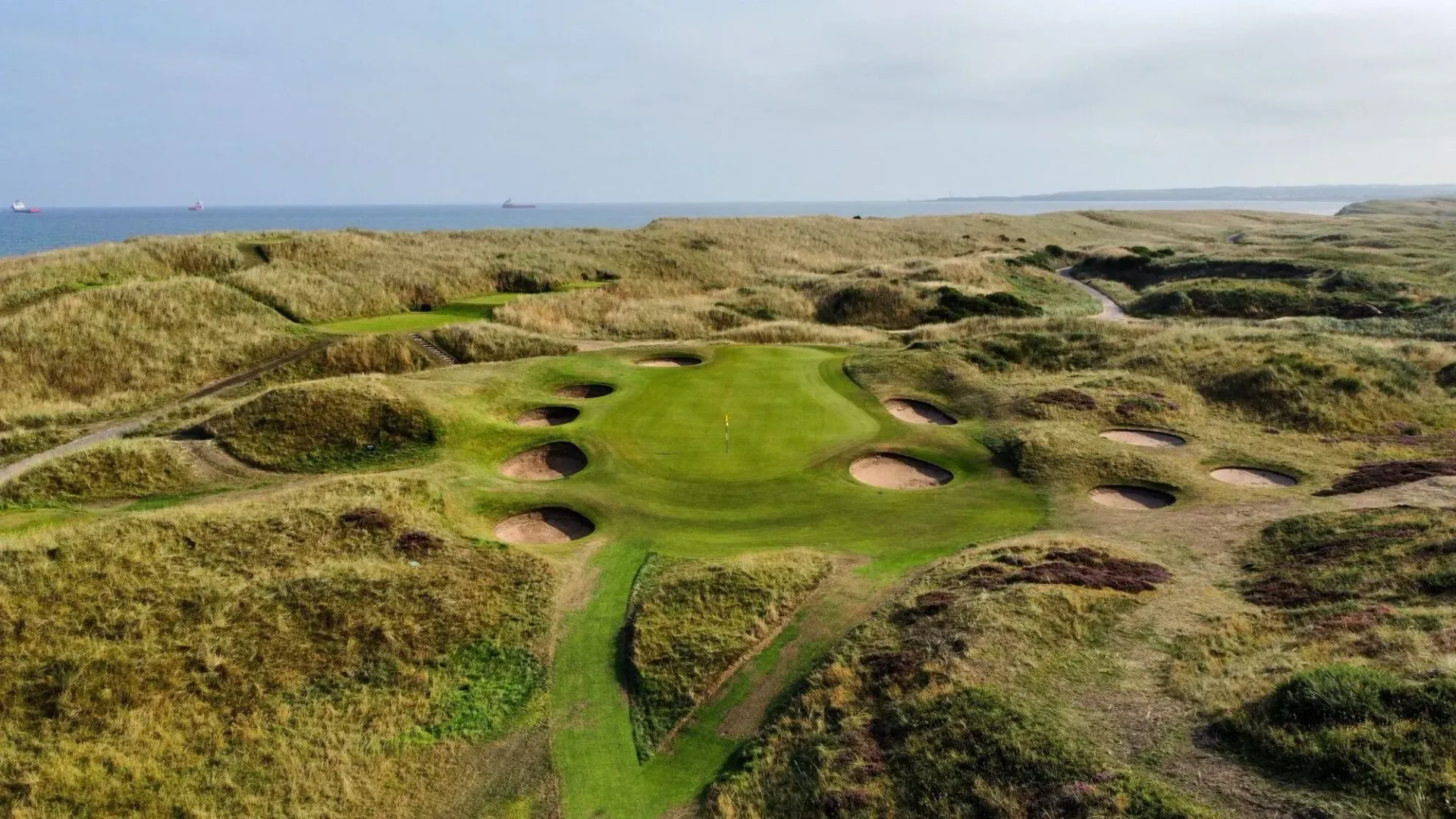 Aerial view of a golf course green surrounded by bunkers and tall, golden grass near the ocean. Royal Aberdeen