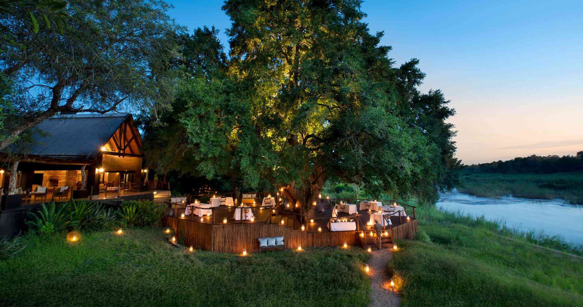 Outdoor dining area with tables under a large tree near a river at dusk, lit by candles. Sabi Sands Game Reserve (Greater Kruger)
