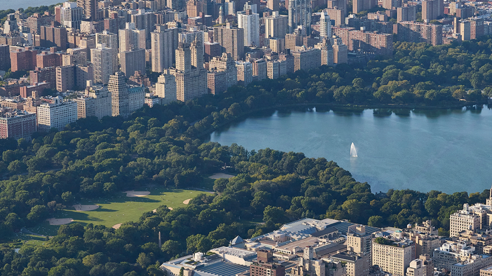 Central Park in NYC, with lake, trees, buildings, and a few boats on the water.