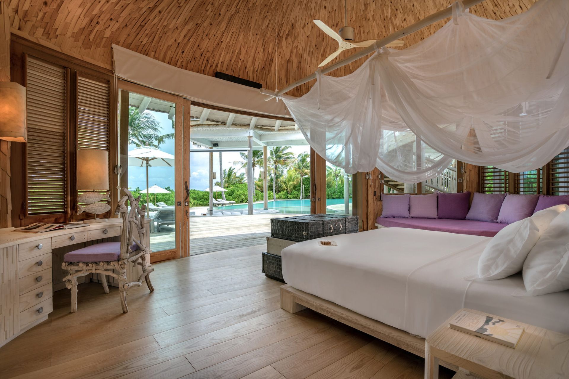 Bedroom with thatch roof, four-poster bed, white furniture, purple couch, and open doors to pool and ocean.The Soneva Jani Resort, Maldives