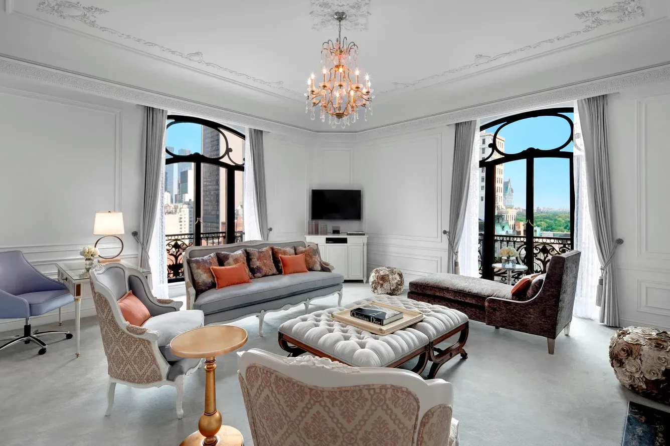 Elegant living room with ornate moldings, chandelier, and city view through arched windows. St. Regis New York hotel suite 