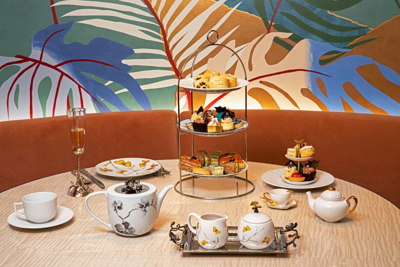 Afternoon tea setting: tiered treats, teapot, teacups, and pastries on a round table, against a tropical mural. St. Regis New York hotel