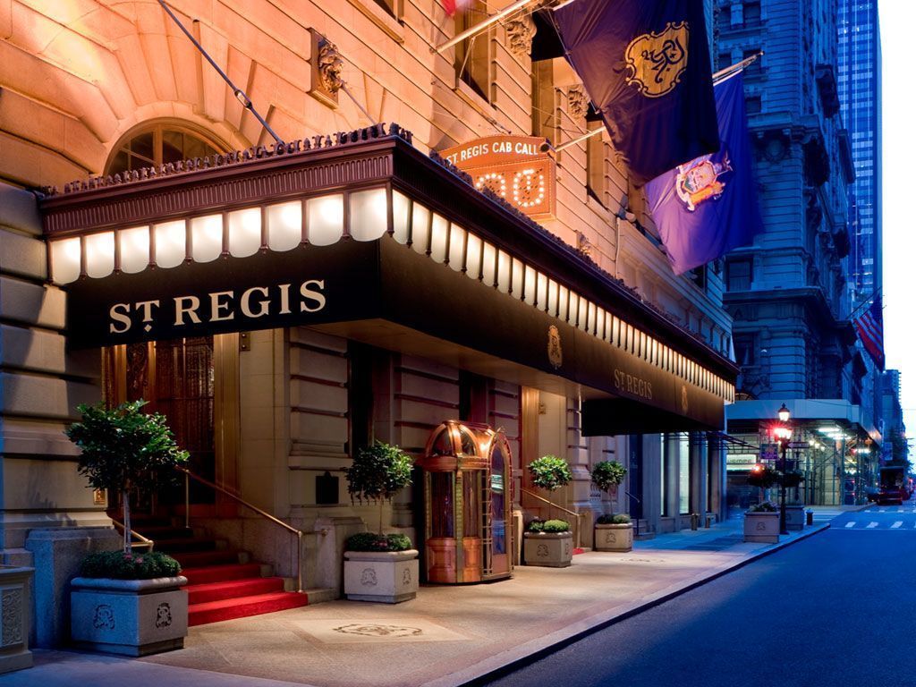 Entrance to the St. Regis hotel with a black awning, red carpet, and flags, lit at dusk.