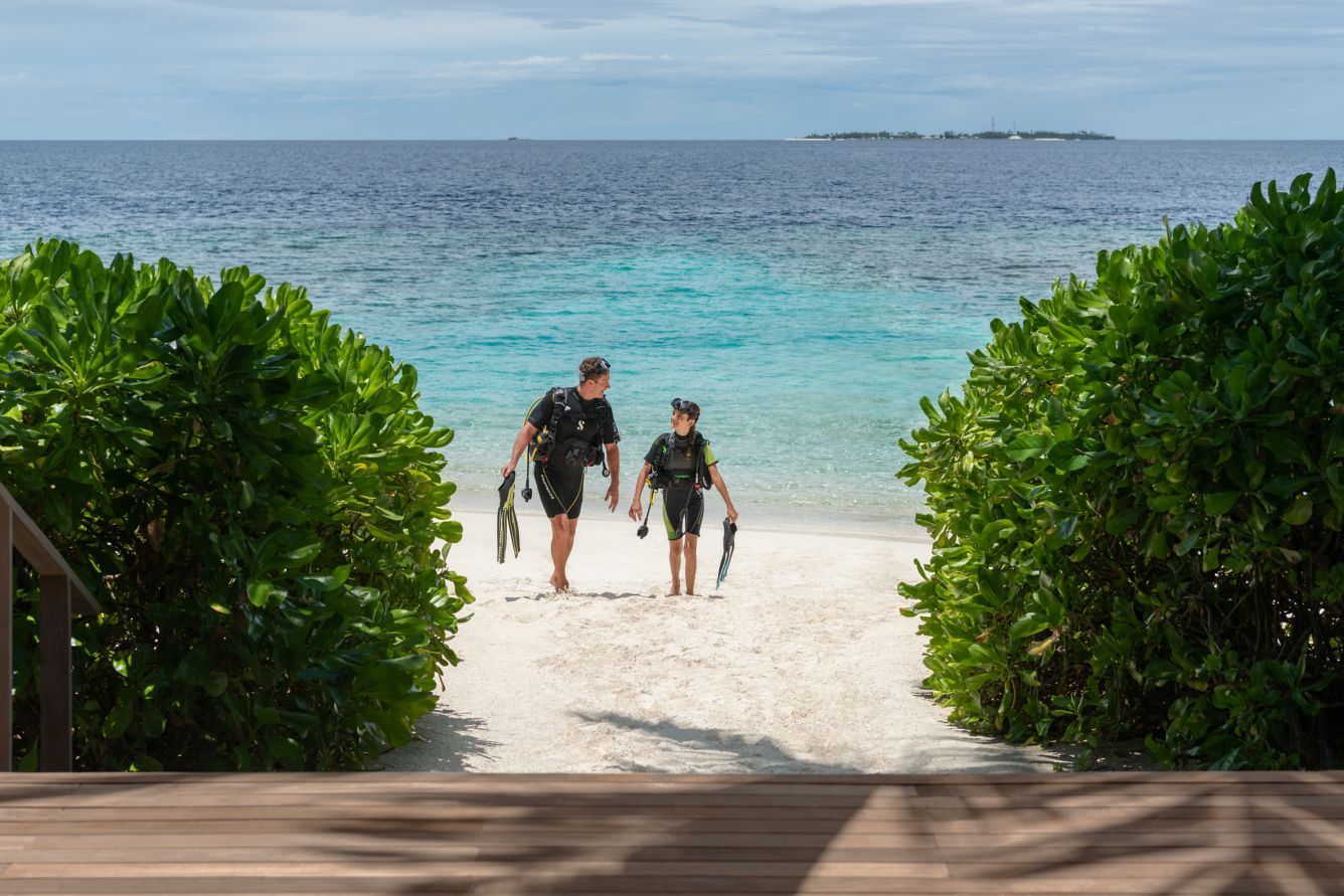 Man and child in scuba gear walk on a white sand beach towards turquoise water, flanked by green bushes.
