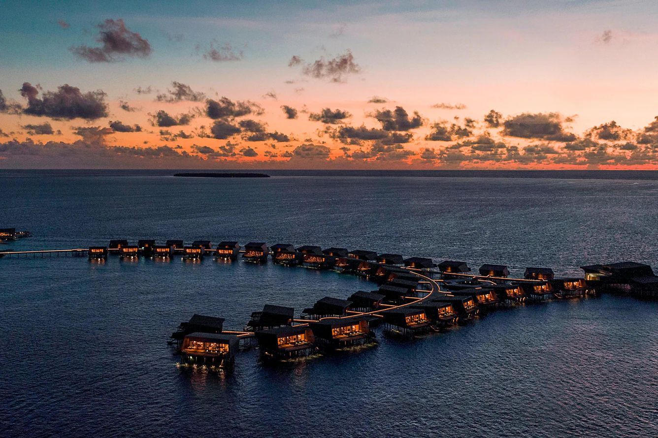 Overwater bungalows at dusk, lit up against a calm ocean with a colorful sunset.