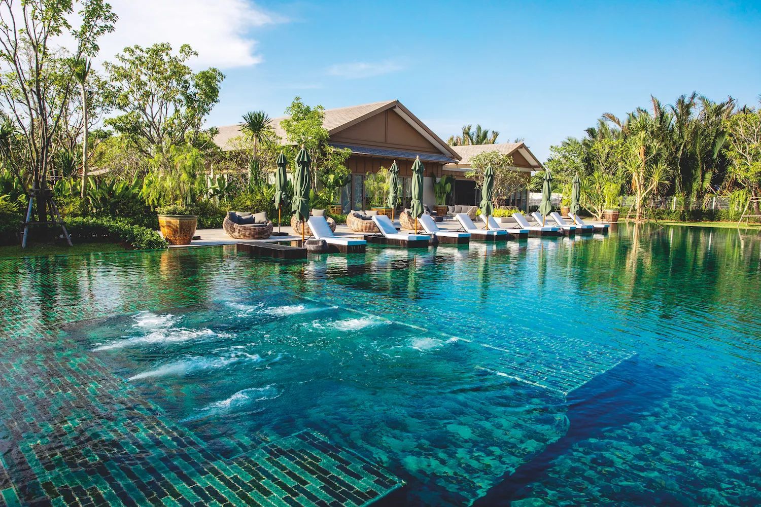 A resort pool with bubbling water in the foreground, facing a row of lounge chairs and a building under a blue sky.