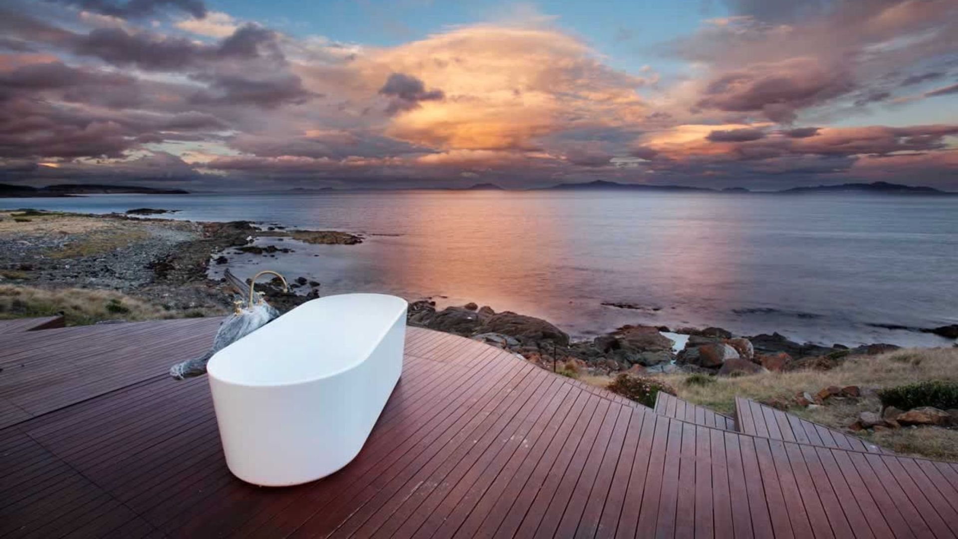 White bathtub on a wooden deck overlooking a calm sea at sunset with colorful clouds.