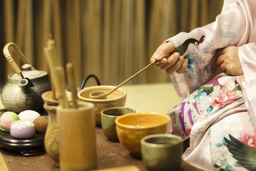 Person in kimono preparing tea ceremony, using a ladle, near pottery and teapot.