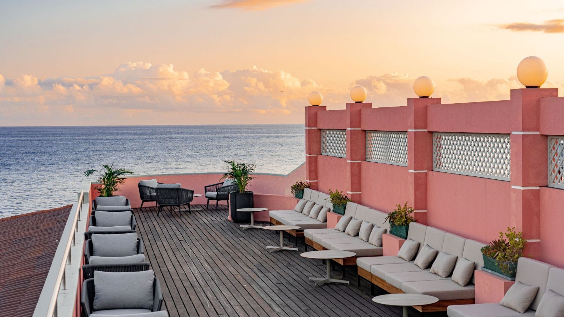 Rooftop patio with ocean view; pink walls, light-colored couches, small tables, and globe lights; setting sun.