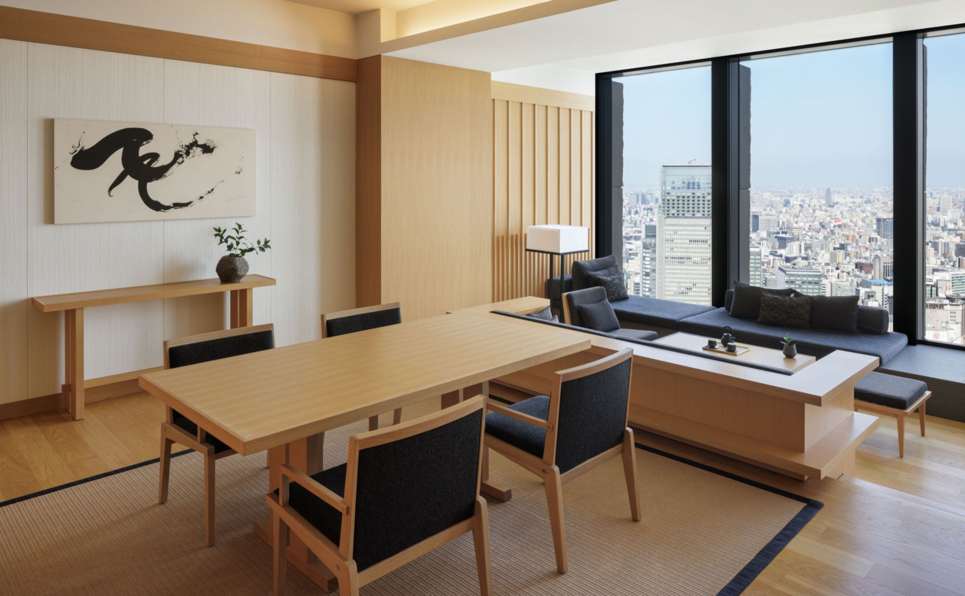 Modern hotel room with dining table, low seating by a large window overlooking a city. Aman Tokyo 