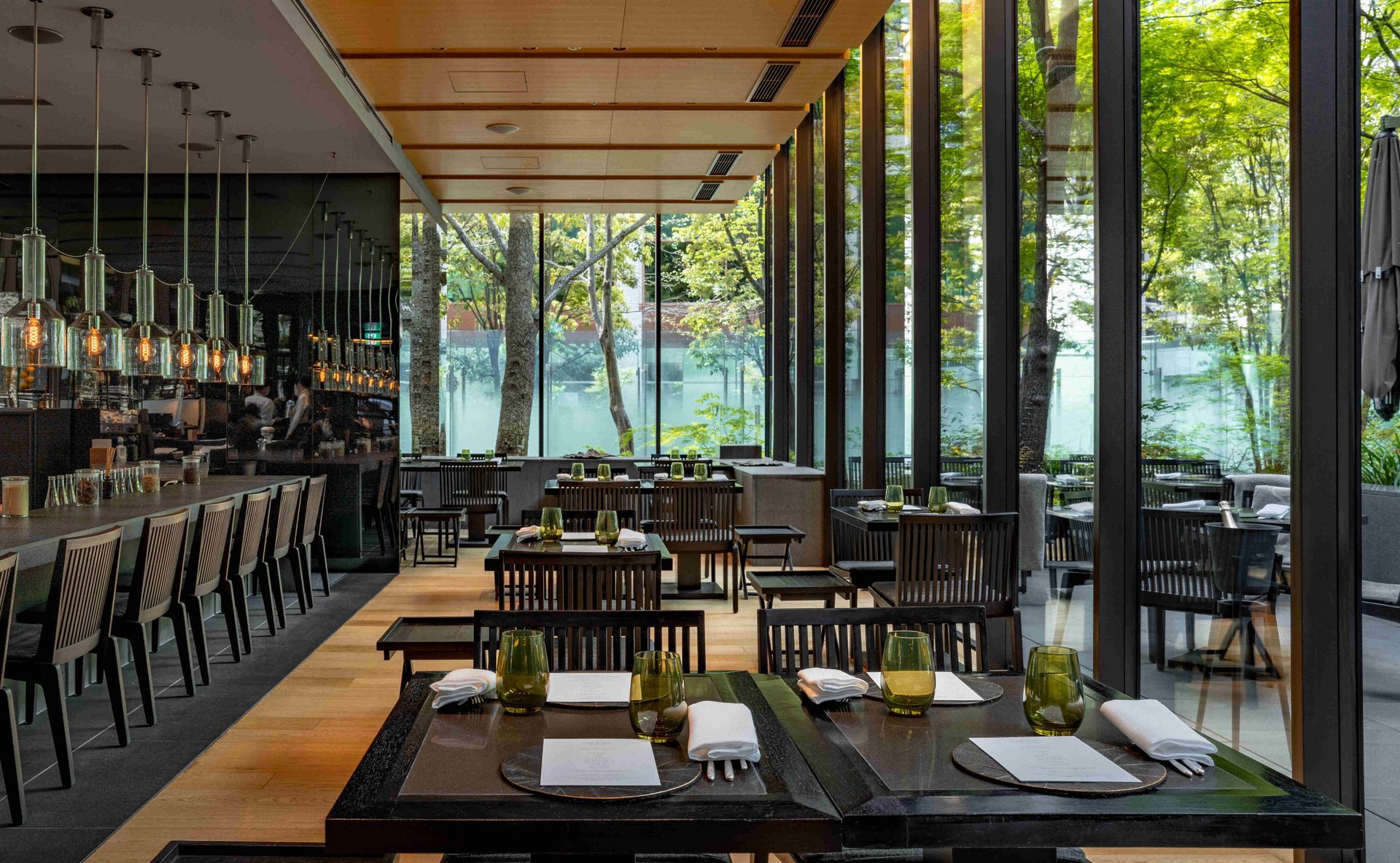 Restaurant interior with tables, chairs, bar, large windows overlooking greenery, and hanging lights. Aman Tokyo 