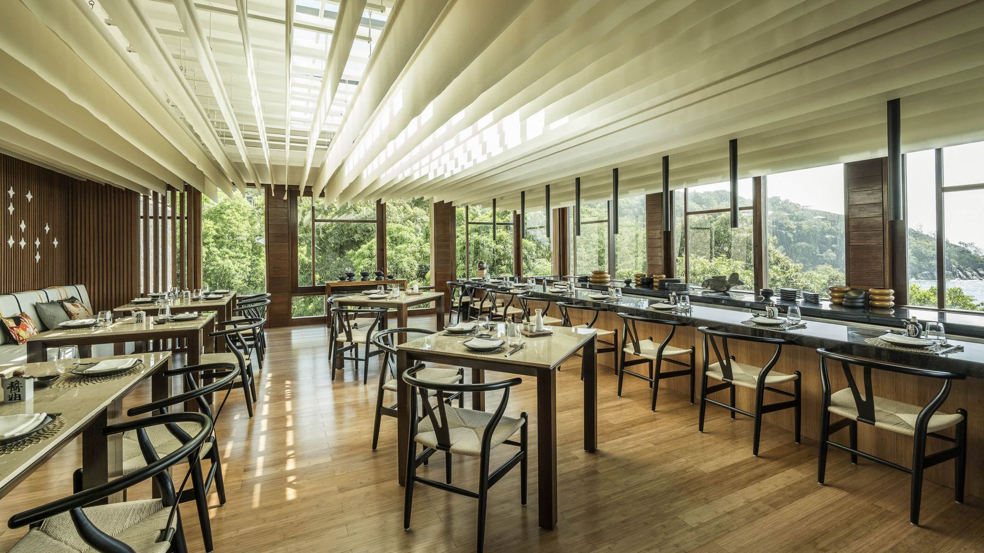 Lush green trees overlooking a blue ocean. Lounge chairs on a wooden deck. Sunny day.