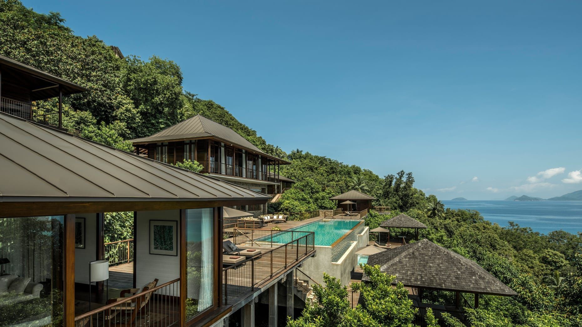 Luxurious hillside resort with pool, ocean view, and lush green foliage under a clear blue sky.