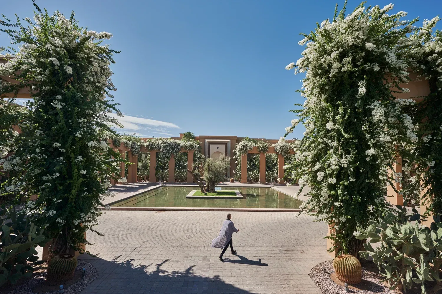 A person walks toward a building framed by flower-covered archways on a sunny day.