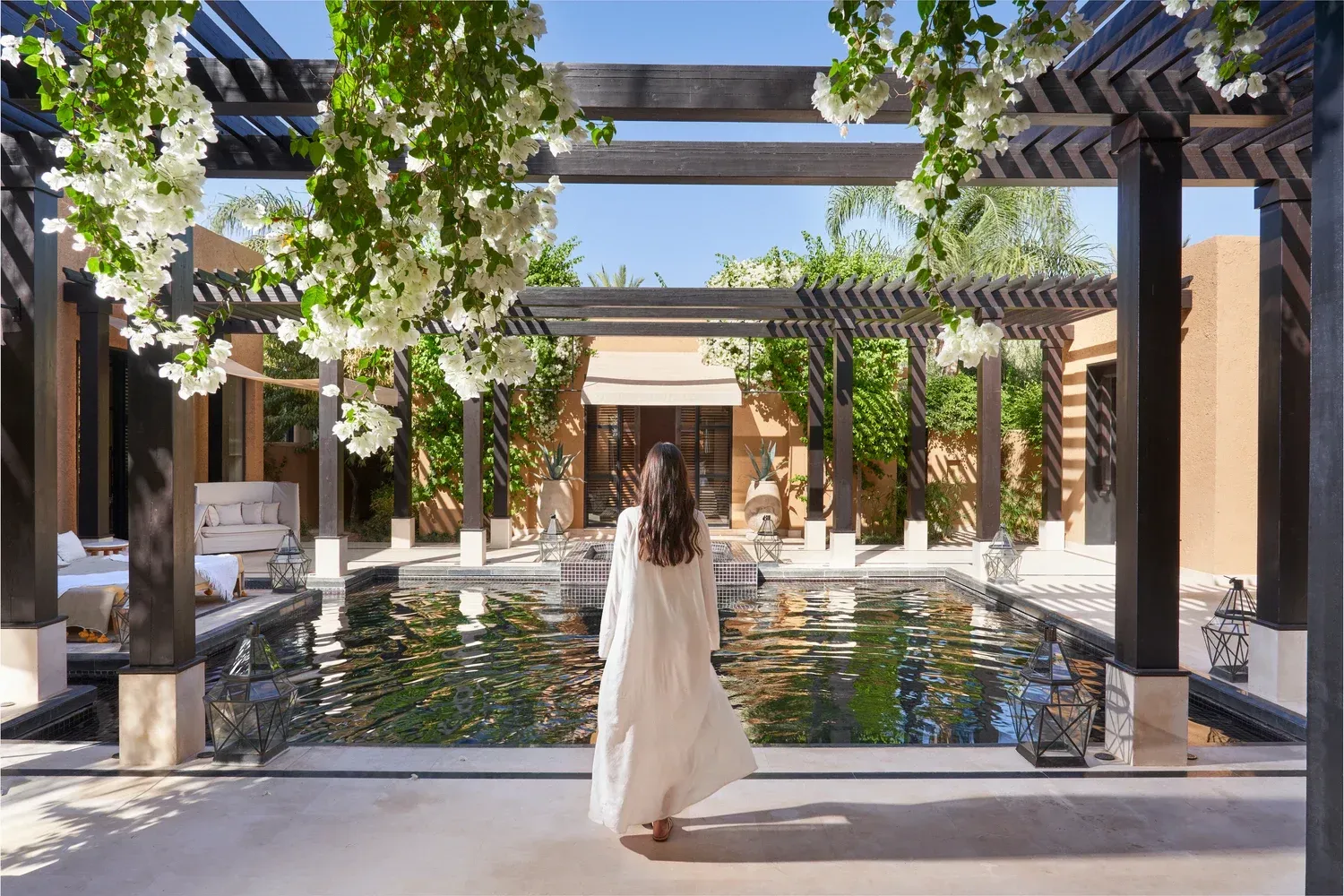 Woman in white robe walks toward a reflecting pool under a pergola draped with white flowers. The Mandarin Oriental, Marrakech