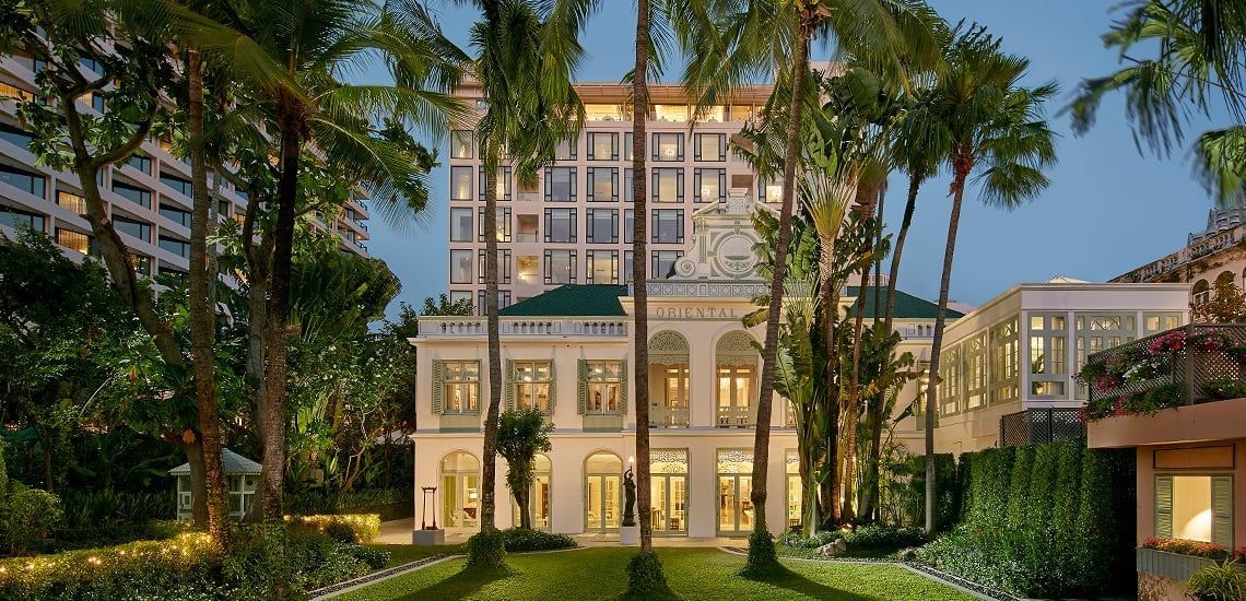 A white colonial building with a modern high-rise in the background, framed by palm trees and lush greenery. The Peninsula Bangkok