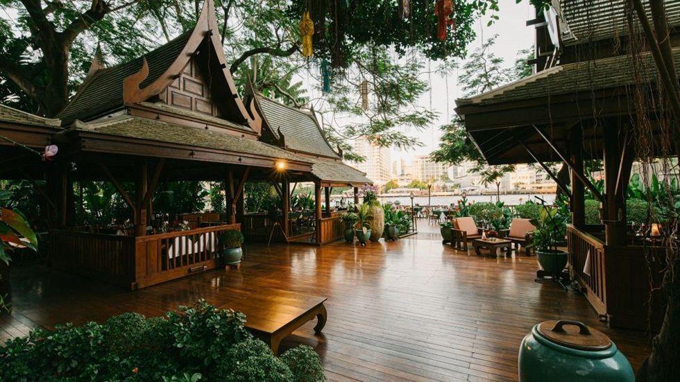 Wooden Thai-style pavilions with intricate roofs, overlooking a waterway with foliage and light. The Peninsula Bangkok