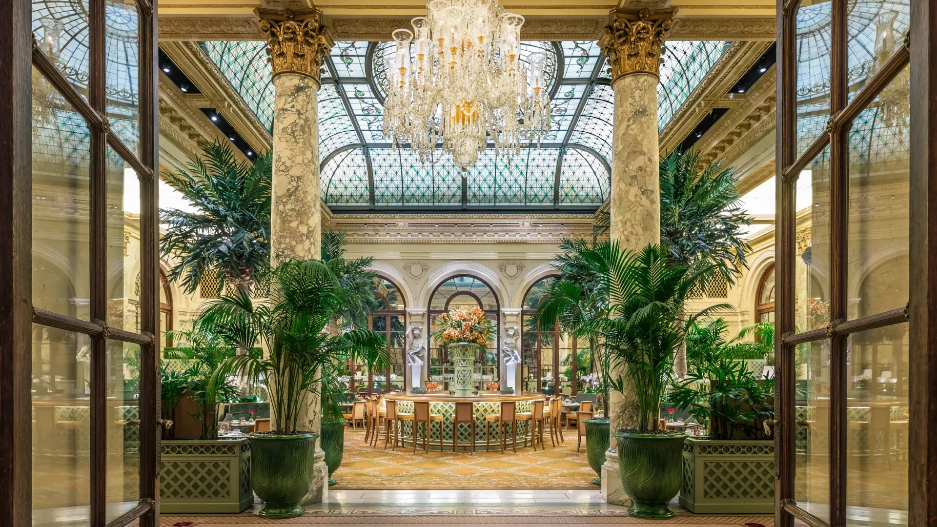 Plaza Hotel New York Elegant hotel dining room, seen through glass doors, features a large chandelier, arched doorways, potted plants, and a central dining table.