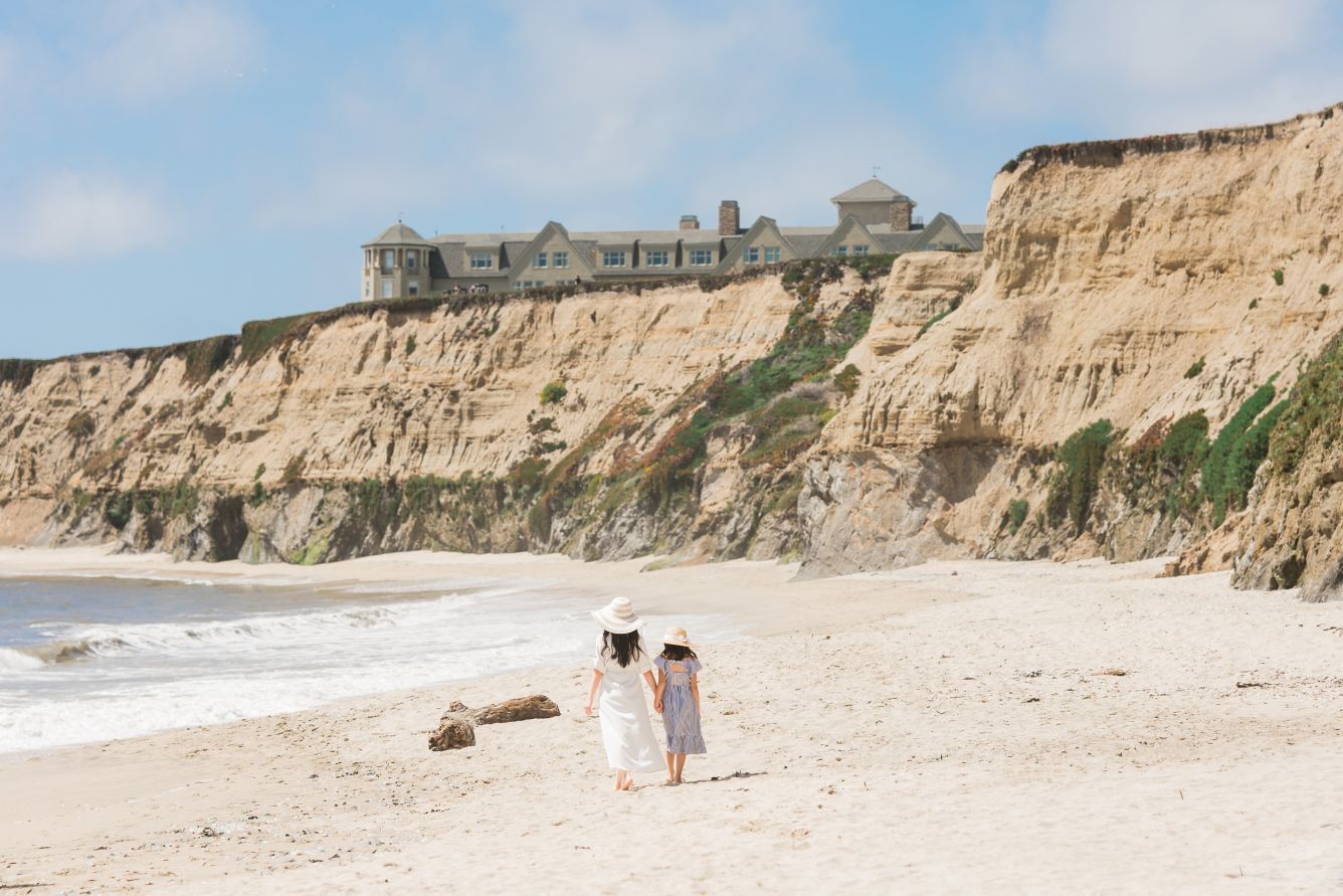 Two people walk on a beach towards a large building on a cliff, sunny day. The Ritz-Carlton, Half Moon Bay, California