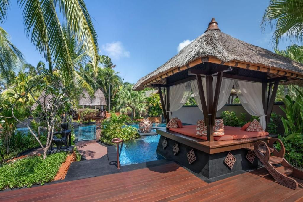 Poolside gazebo with cushions, curtains, and thatched roof; tropical setting with palm trees. The St. Regis Bali Resort