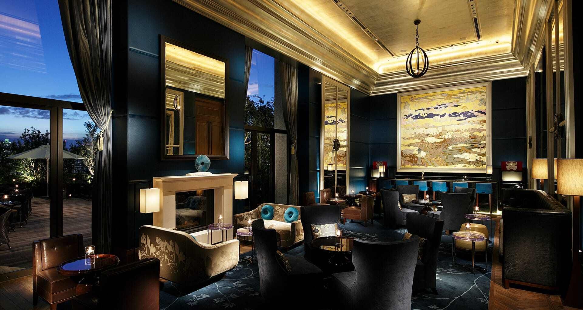 A luxurious lounge with dark blue walls, a fireplace, and a large artwork, with an outdoor terrace visible through large windows. The St. Regis Osaka