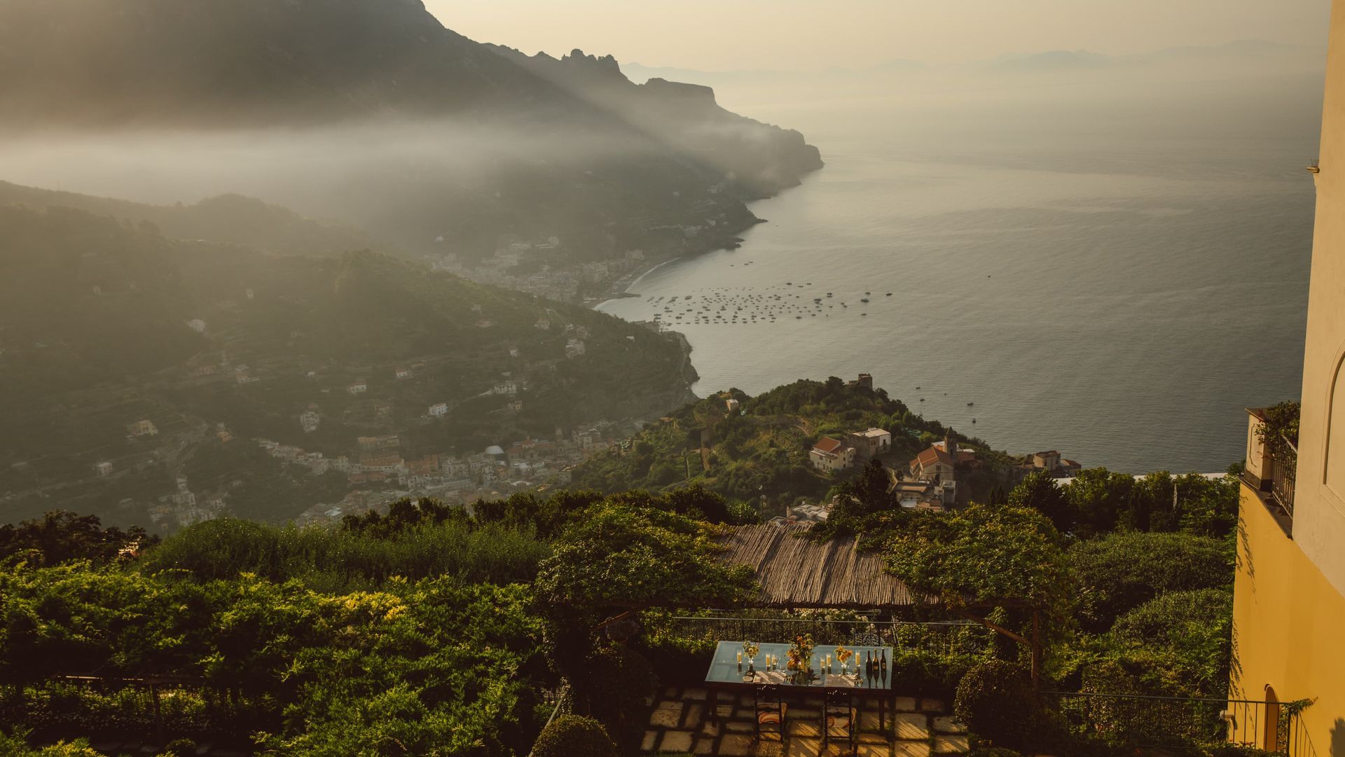 Coastal view of Italian mountains and sea in hazy sunlight. Green foliage in foreground, buildings along the coast. The Belmond Hotel Caruso, Ravello