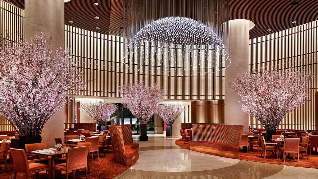 The Peninsula Tokyo A luxurious restaurant lobby with pink flowering trees, orange seating, and a large, round light fixture.