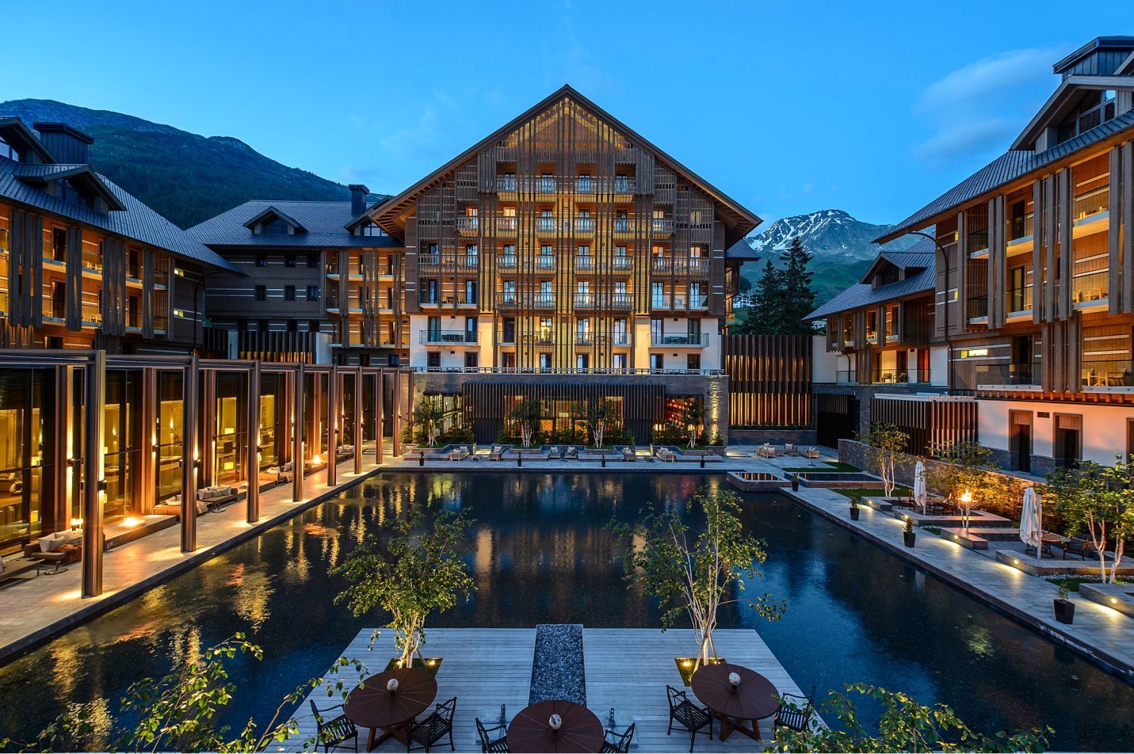 Luxury hotel with wooden exterior, pool, and mountain backdrop at dusk. The Chedi Andermatt_