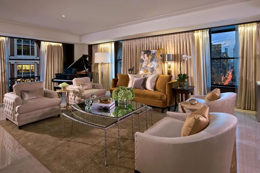 Elegant living room with a grand piano, velvet sofa, armchairs, and floor-to-ceiling windows with drapes.  The Peninsula hotel on 5th Avenue, NYC