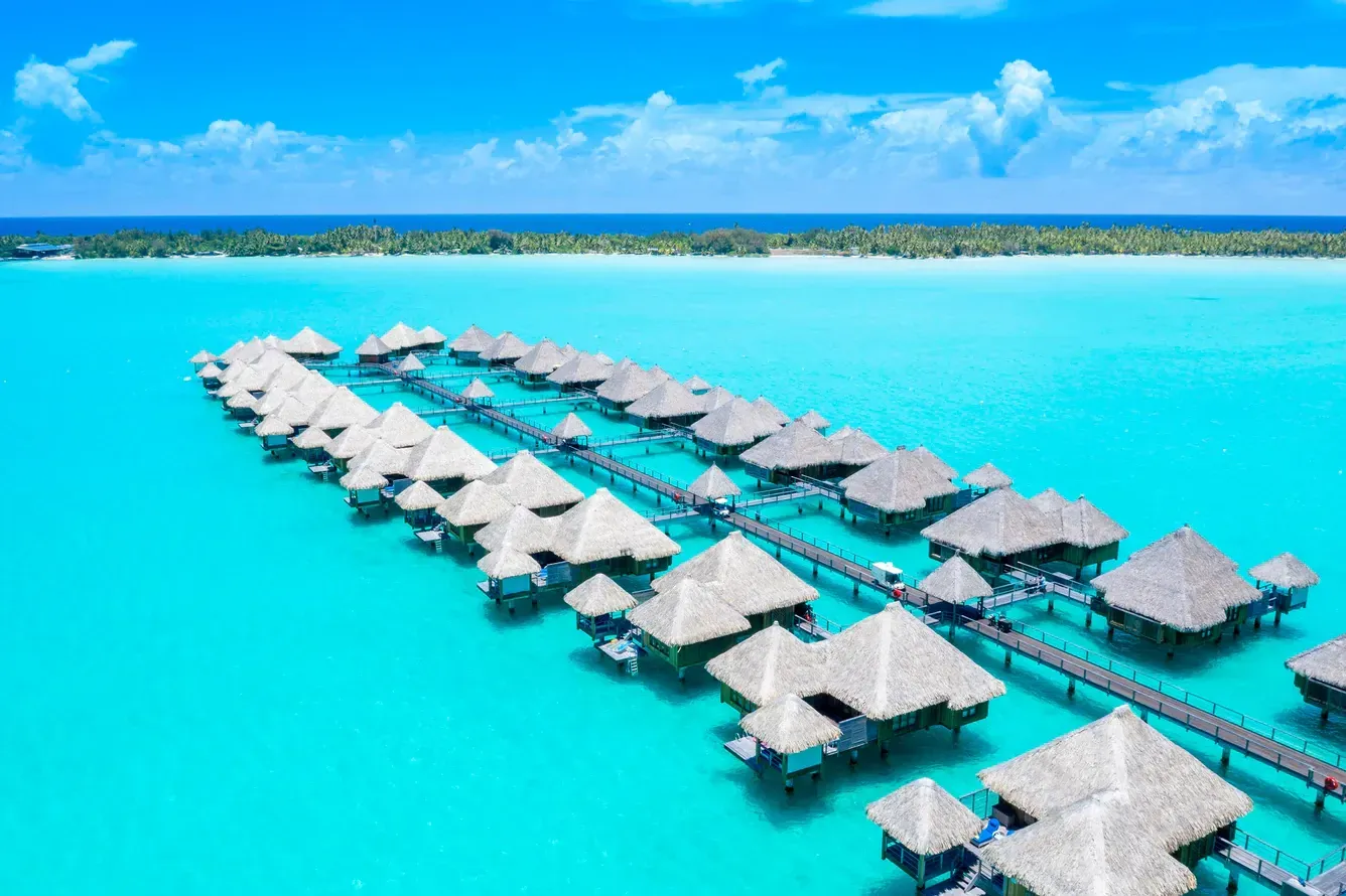 Overwater bungalows in turquoise water; tropical resort with thatched roofs, blue sky, and a distant island. The St. Regis, Bora Bora