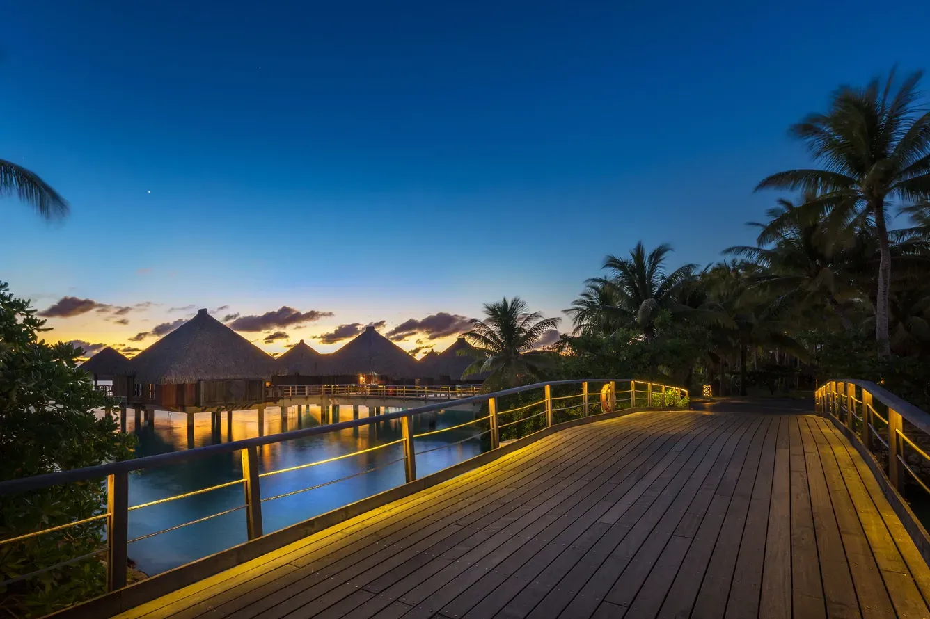 Wooden walkway lit with yellow lights leads to overwater bungalows at dusk. The St. Regis, Bora Bora