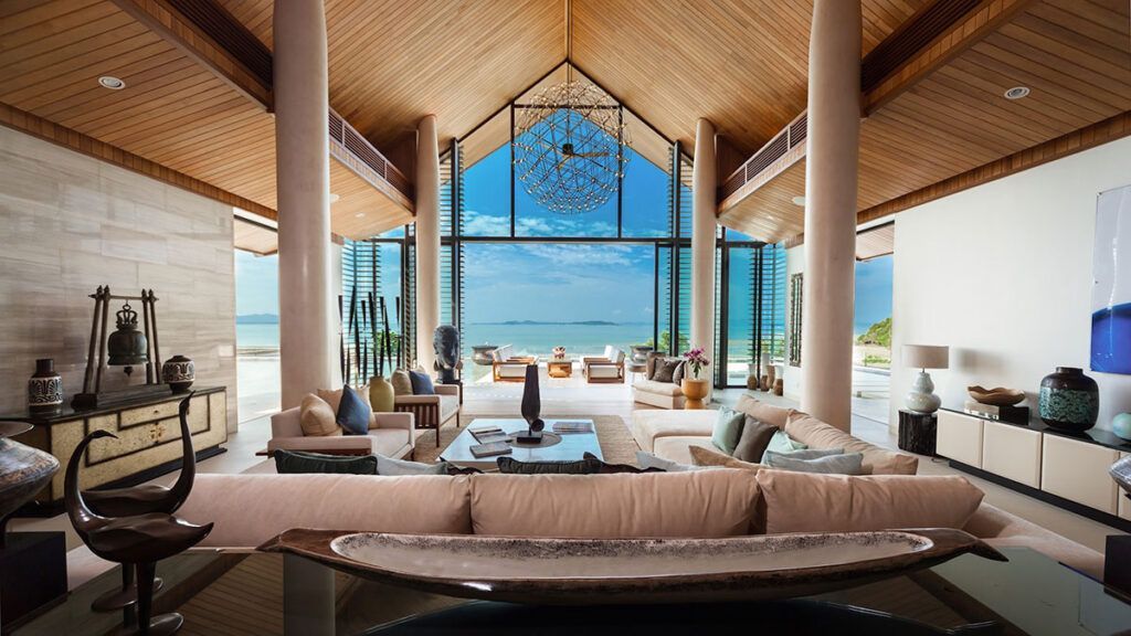 Luxurious beachside living room with ocean view. Beige and brown tones, floor-to-ceiling windows, and modern furniture. Villa Amarapura – Phuket