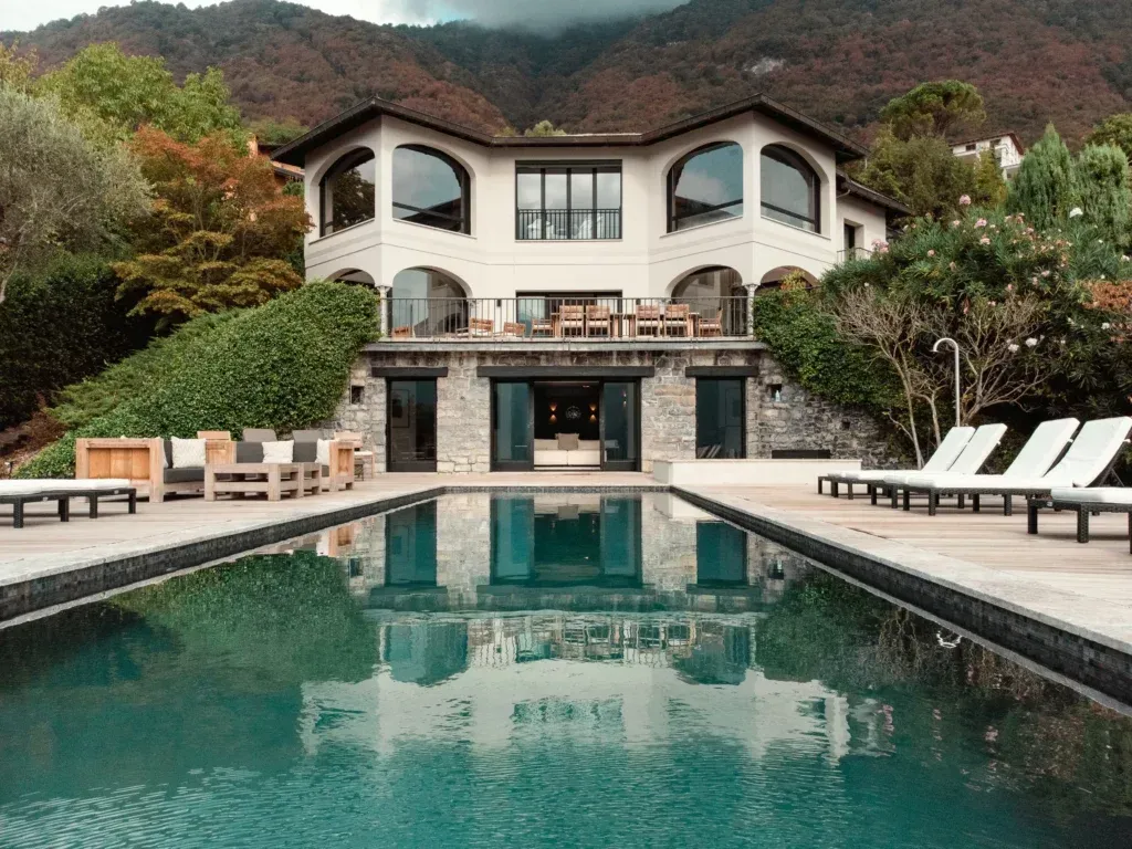 A white multi-story villa with arched windows sits behind an infinity pool against a backdrop of mountains.