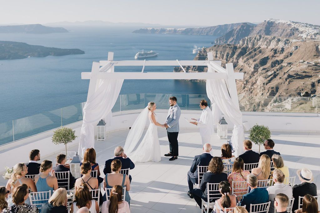 A couple stands under a white wedding arch on a cliffside terrace in Santorini overlooking the sea, with guests seated.