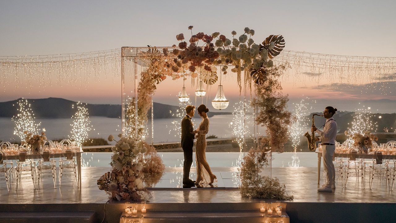 A couple stands under a flower-adorned wedding arch at sunset, overlooking a calm ocean with shimmering, sparkling lights.