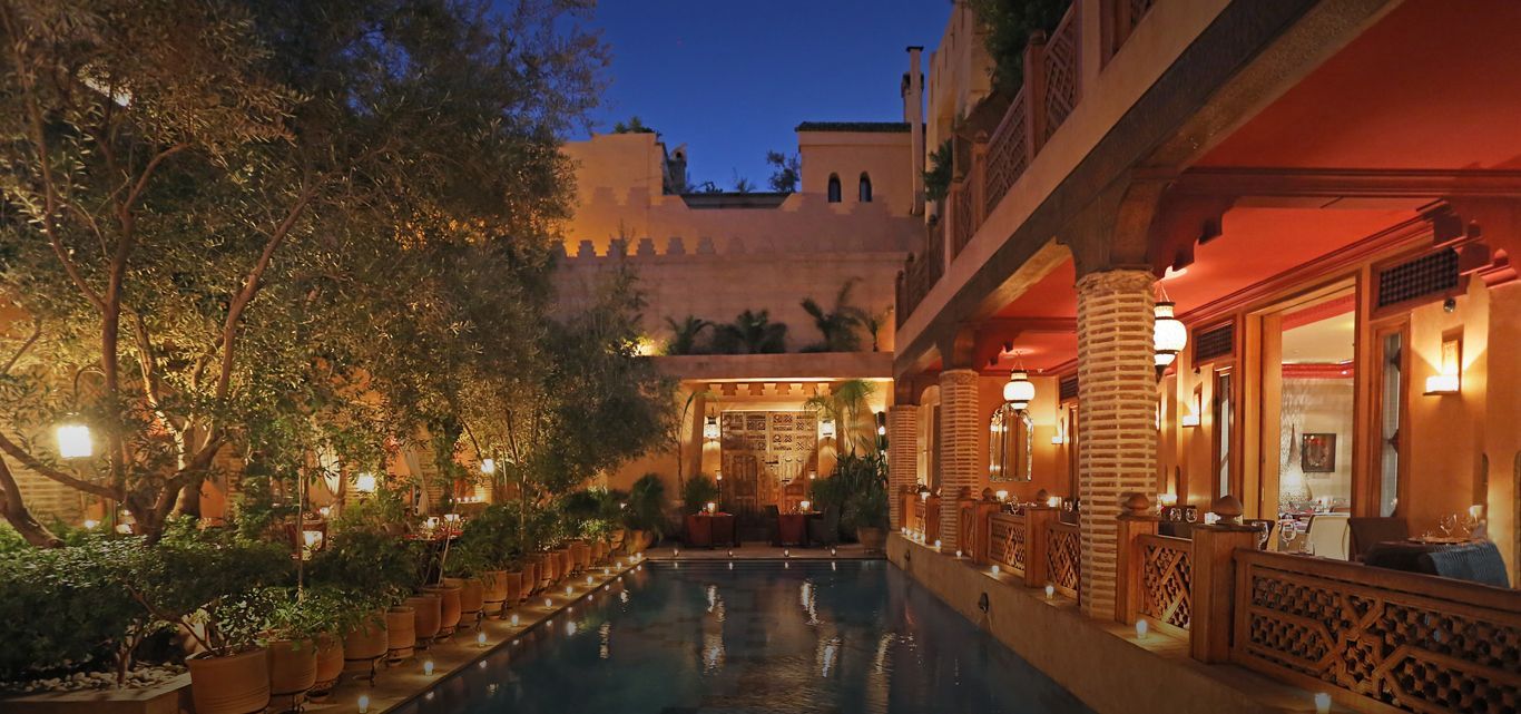 An outdoor hotel pool courtyard at dusk, featuring warm lighting, lush greenery, and traditional architectural details.