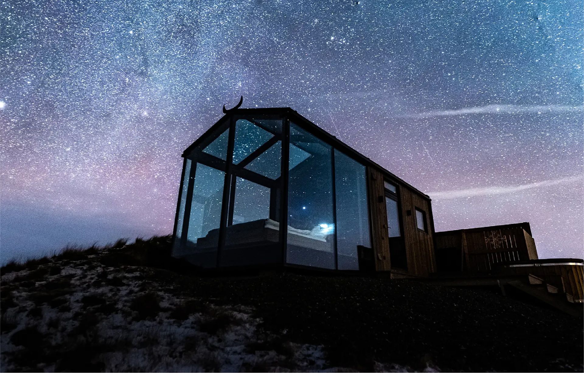 A modern glass-walled cabin sits on a dark hillside at night under a vibrant, star-filled sky. ÆRA Panorama Glass Lodge