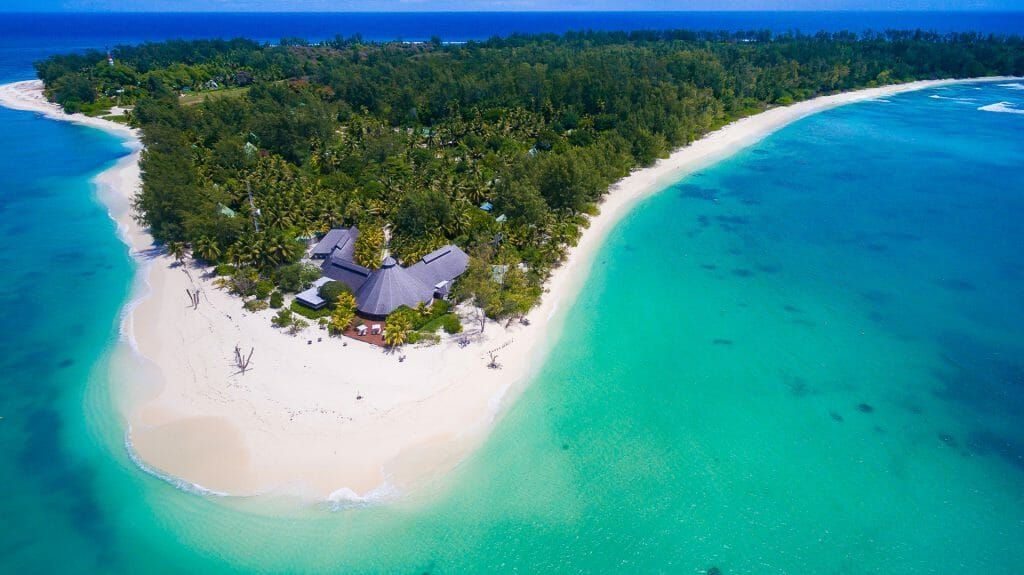 Aerial view of a tropical island with a thatched-roof resort, white sandy beach, and clear turquoise water.