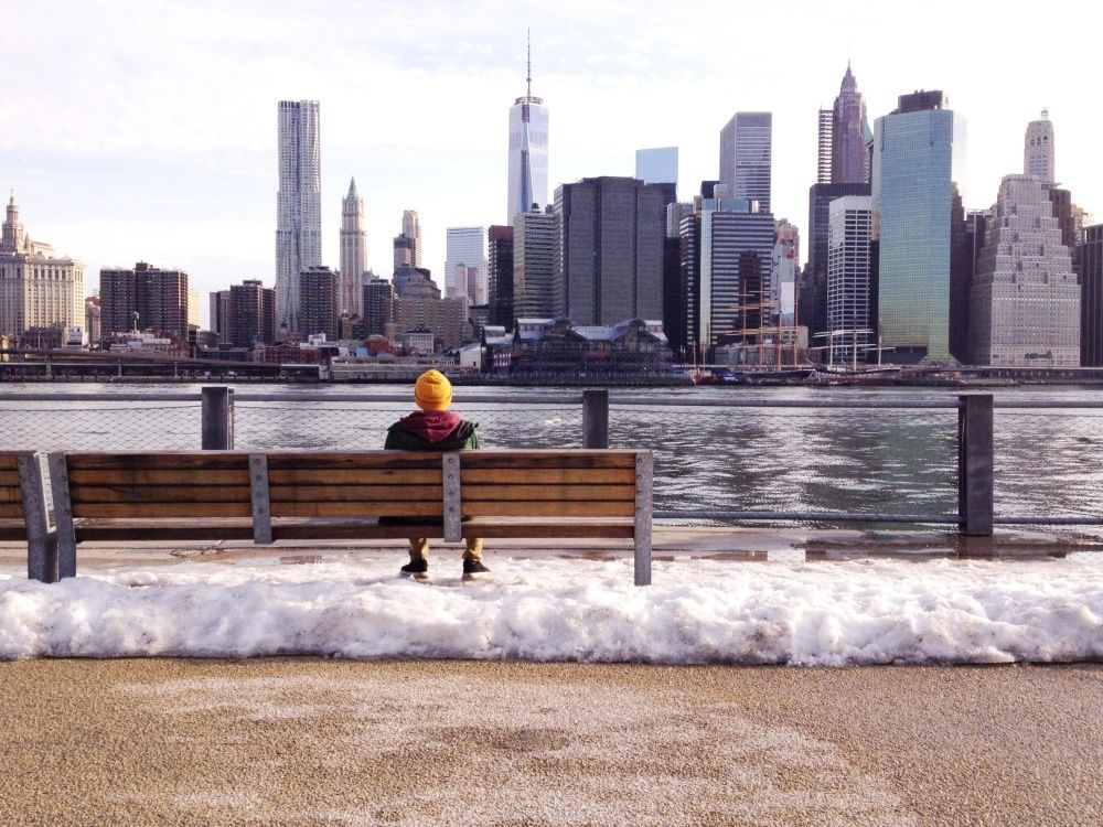 Person sits on a bench overlooking the New York City skyline with snow in foreground.