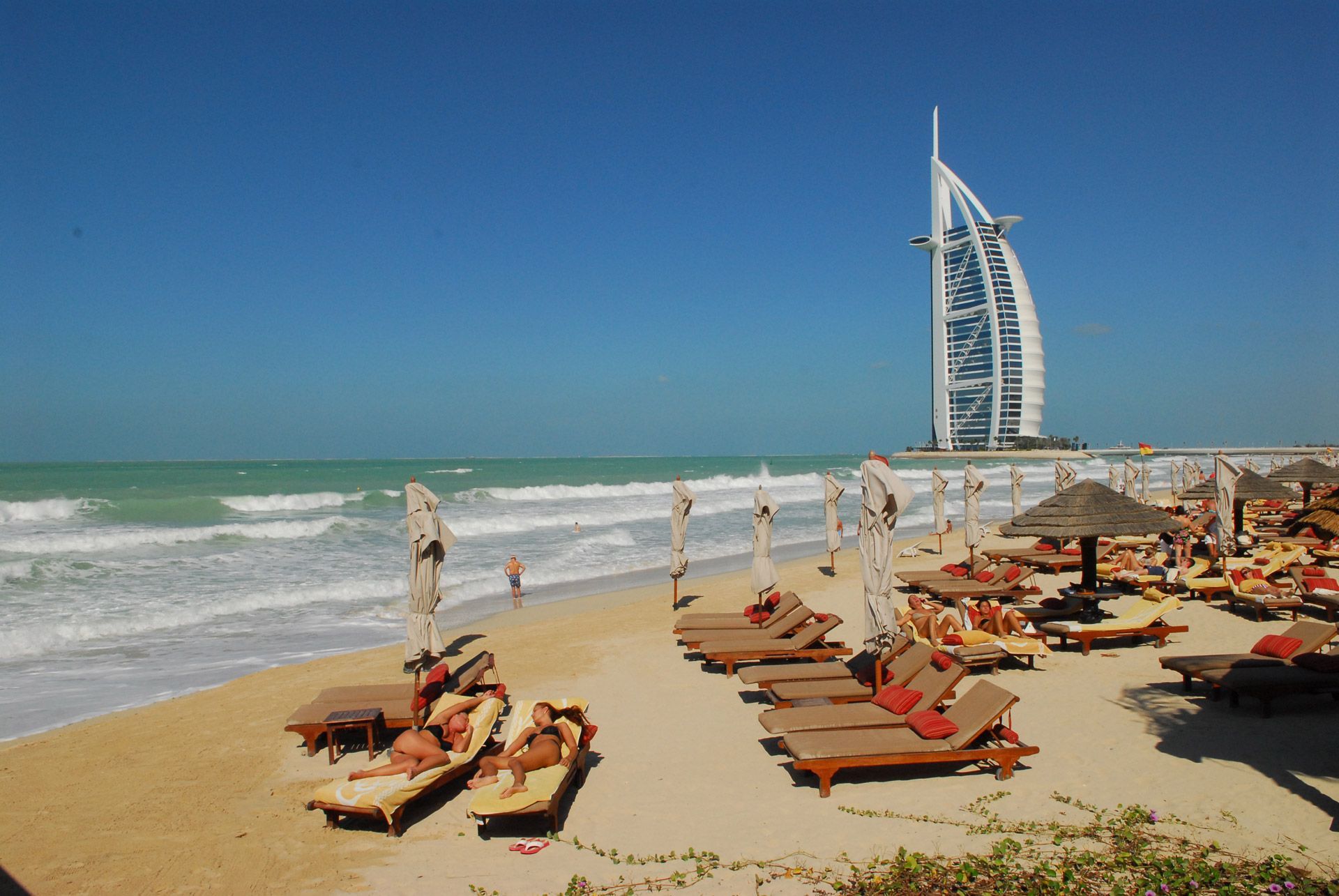 Beach with lounge chairs, ocean, and the Burj Al Arab hotel in Dubai under a blue sky.