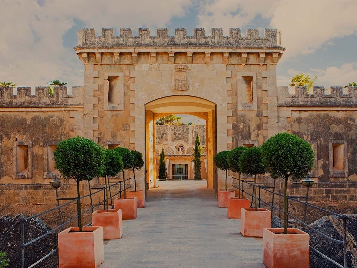 A stone fortress entrance with a symmetrical row of potted topiary trees lining the walkway leading to the archway.