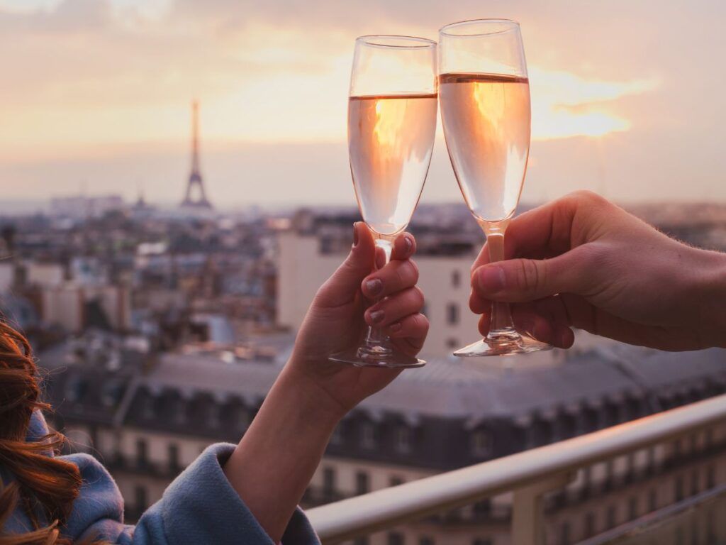 Cheers! Two hands holding champagne flutes, toasting with the Eiffel Tower in the background at sunset.